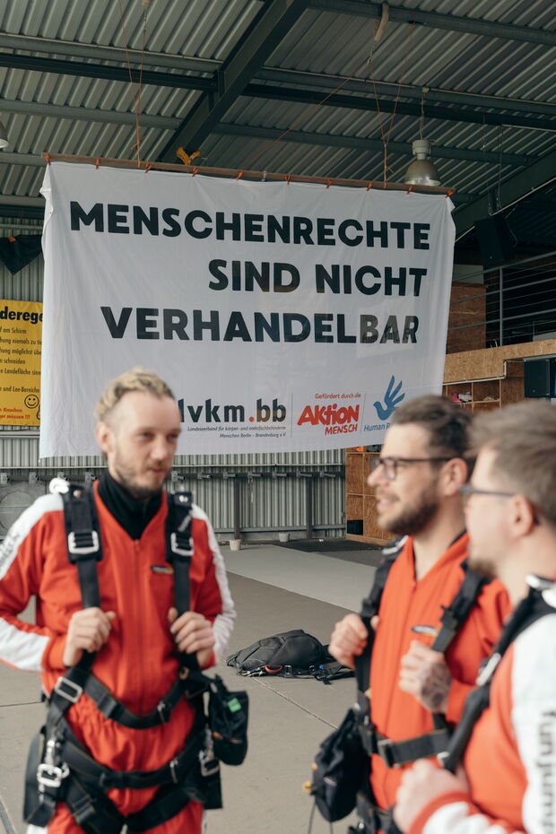 Drei Fallschirmspringer in orange-schwarzer Ausrüstung stehen vor einem Banner mit der Aufschrift „Menschenrechte sind nicht verhandelbar“.