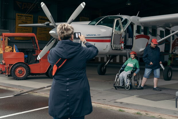 In einer Flugzeughalle fotografiert eine Frau eine Person im Rollstuhl und einen Mann vor einem kleinen Flugzeug. Im Hintergrund steht ein roter Gabelstapler.