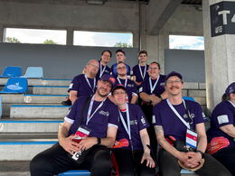 Ein Gruppe von Sportlern in lila Shirts sitzen und stehen auf einer Tribüne, sie lächeln in die Kamera. Hintergrund: Betonpfeiler, blauer Himmel