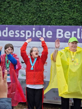 Madlen und zwei weitere triumphierende Athlet:innen mit Medaillen und Regenponchos feiern ihren Sieg auf dem Podium.