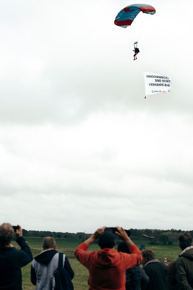 Eine Gruppe Menschen beobachtet einen Fallschirmspringer mit Banner „Menschenrecht sind nicht verhandelbar“ am bewölkten Himmel.