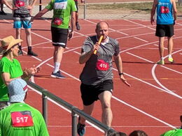 Staffelläufer Michael in grauer Sportkleidung mit Startnummer 416 übergibt Staffelstab auf roter Laufbahn, Hintergrund: andere Läufer, Zuschauer, Bäume