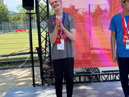 Sieger Jordan in grauer Sportkleidung mit Medaille applaudiert auf roter Bühne mit Zahl 8, Hintergrund: großer Bildschirm, Zaun, Bäume