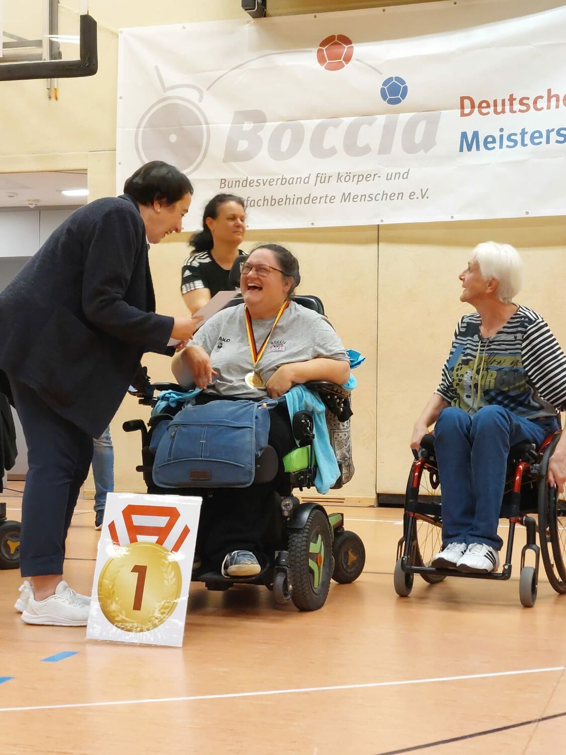 Rollstuhlfahrer mit Medaille bei Boccia-Meisterschaft, Frau gratuliert.