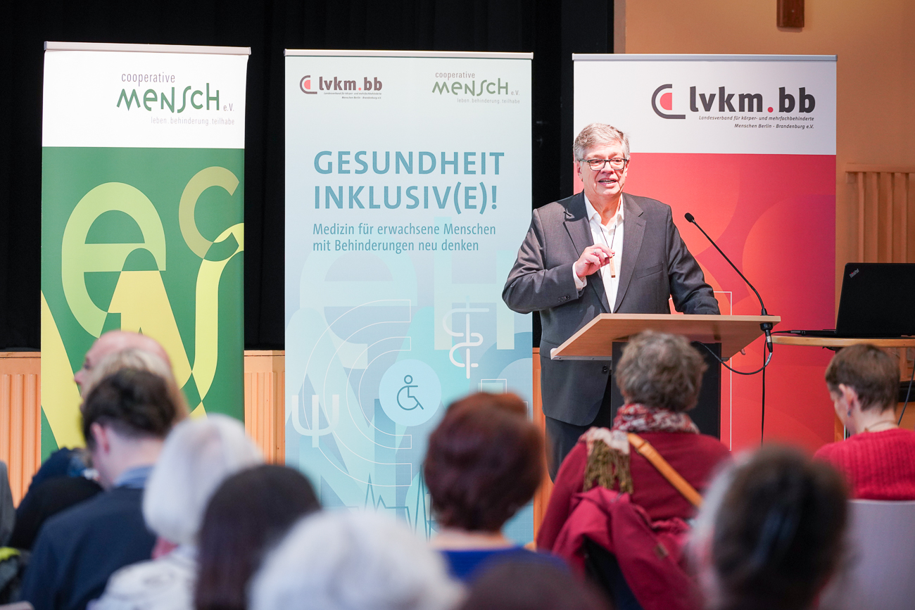 Ein Mann hält eine Rede vor einem Publikum, umgeben von Bannern mit Gesundheitsinformationen.