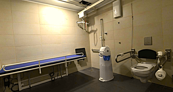 Barrierefreies WC mit höhenverstellbarer Liege, Haltegriffen, Deckenlifter und modernem Toilettensystem. Der Raum ist hell, funktional und großzügig gestaltet, ausgestattet für Menschen mit hohem Pflegebedarf oder Assistenzbedarf.